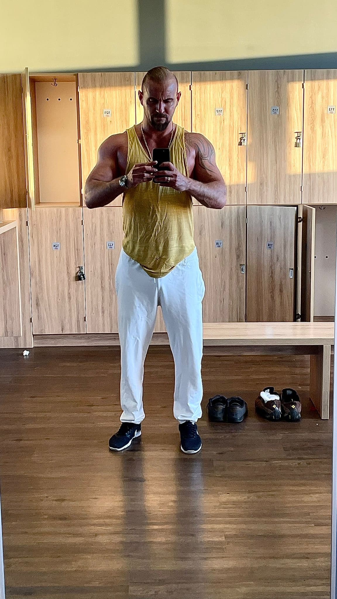 Muskulöser Mann im gelben Tanktop macht ein Spiegelselfie in einer hellen Umkleidekabine vor Holzschränken.