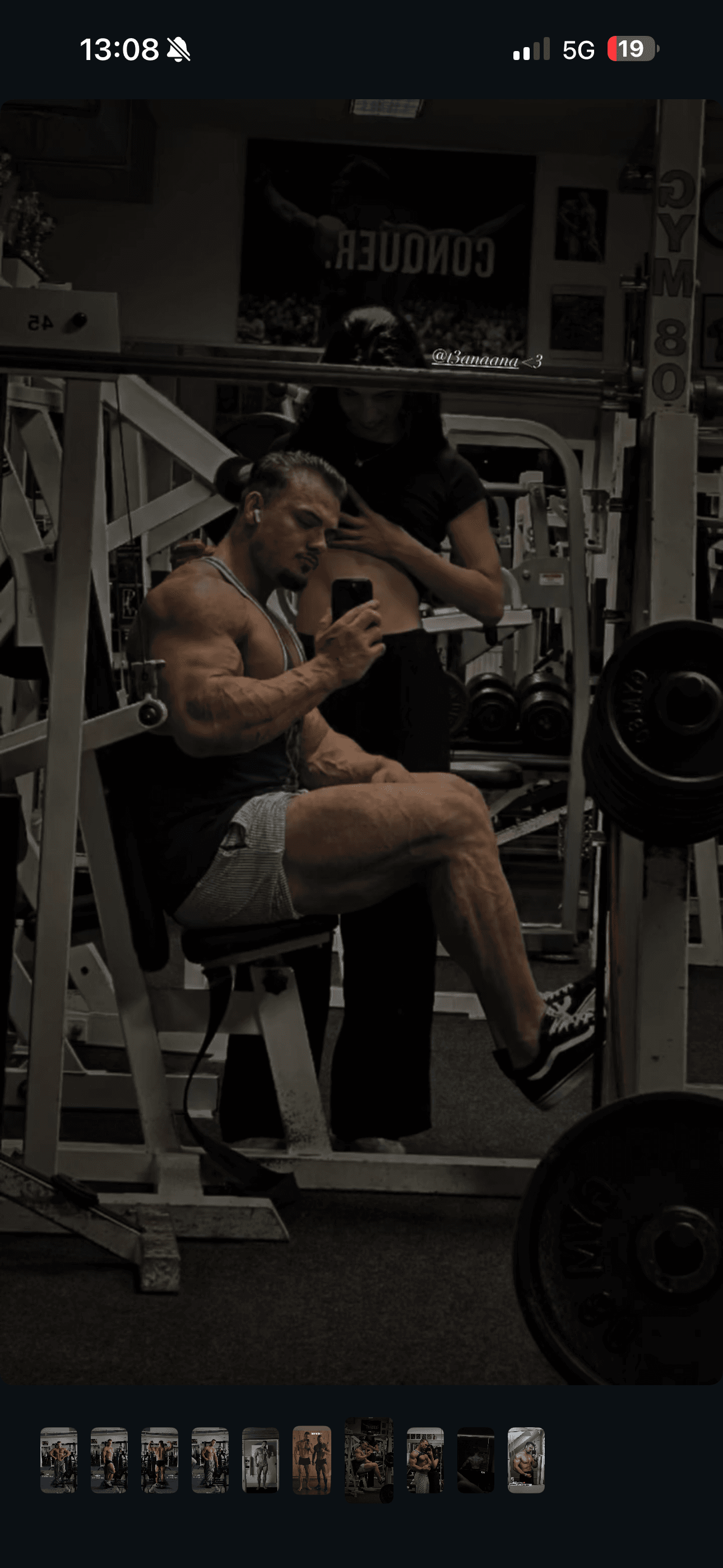 Muskulöser Mann macht ein Spiegelselfie an einem Fitnessgerät, eine Frau steht lächelnd hinter ihm.