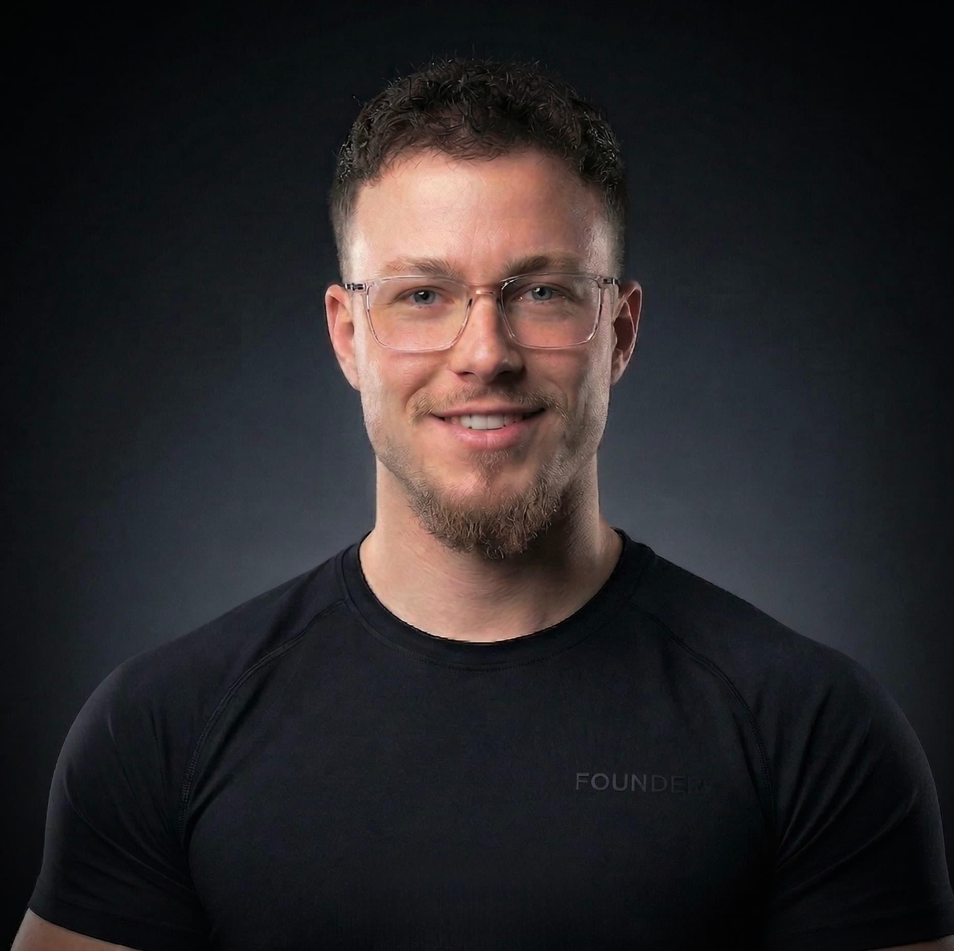 Lächelnder Mann mit Brille und Ziegenbart trägt ein schwarzes T-Shirt vor einem dunklen Hintergrund.