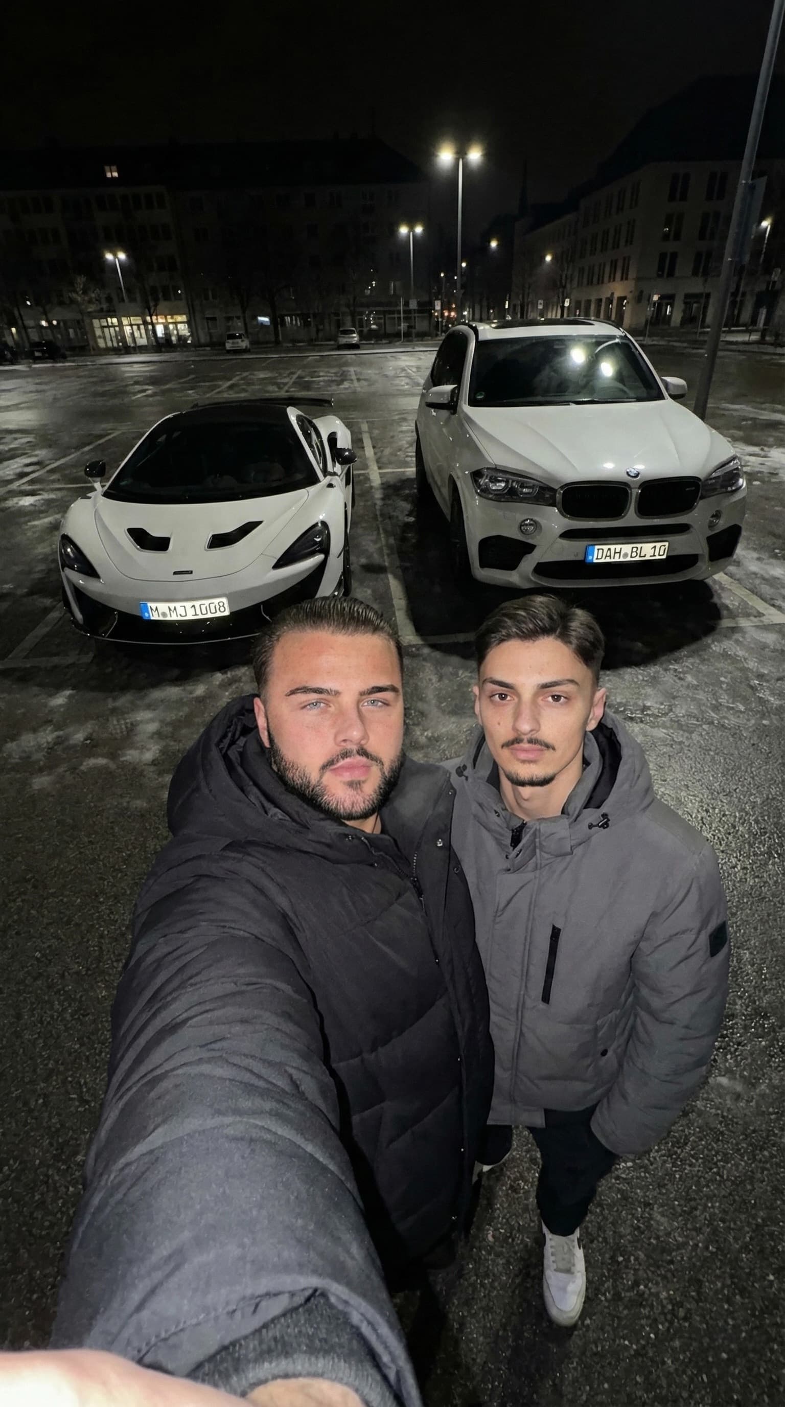 Zwei junge Männer machen nachts ein Selfie vor einem weißen McLaren und einem weißen BMW.